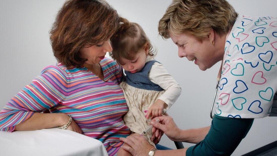 A health professional gives a shot to a young girl who is being held by her mother